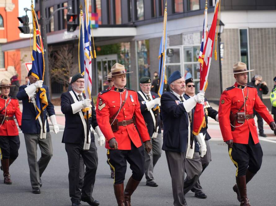 RCMP officers in red serge marching in a parade while carrying flags during a public ceremony in Canada.